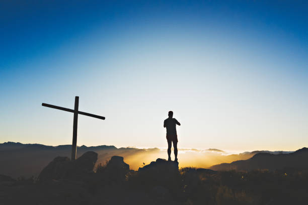 One male trail runner hiker silhouette standing on a rock next to a cross and cairn mountain beacon on a mountain summit at sunrise, Simonsberg, Stellenbosch, Cape Winelands, Western Cape, South Africa.